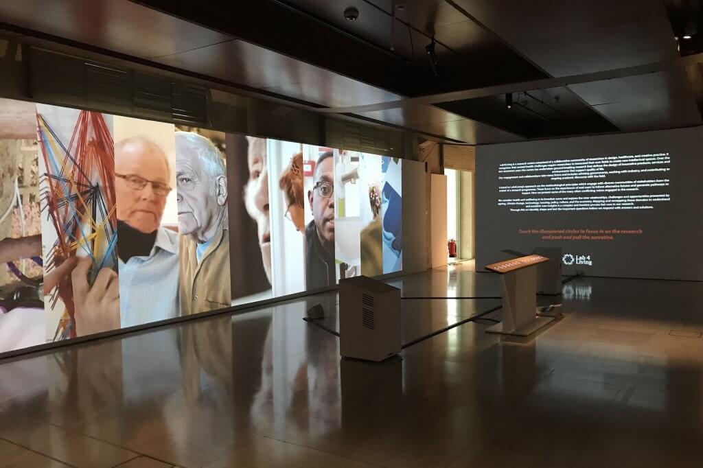 View of the exhibition includes images projects onto a wall (left), descriptive text on the back wall, and the visitor touch panel in the middle of the hall.