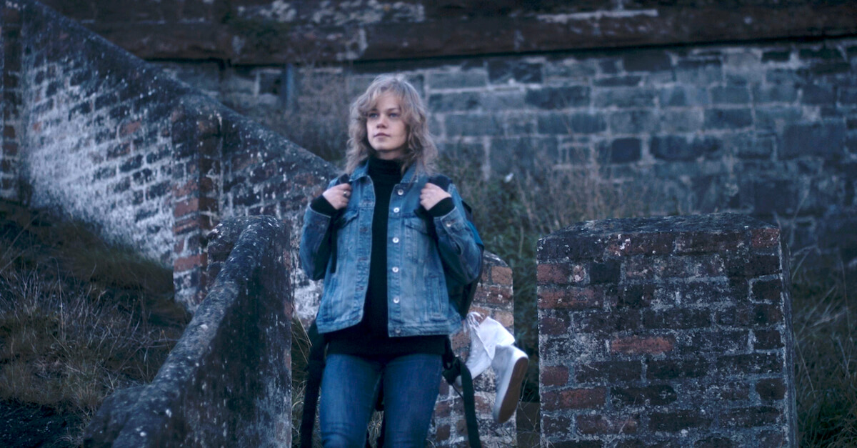 Actress Greta Trusina playing Inga Maria Hauser, standing on steps with back pack