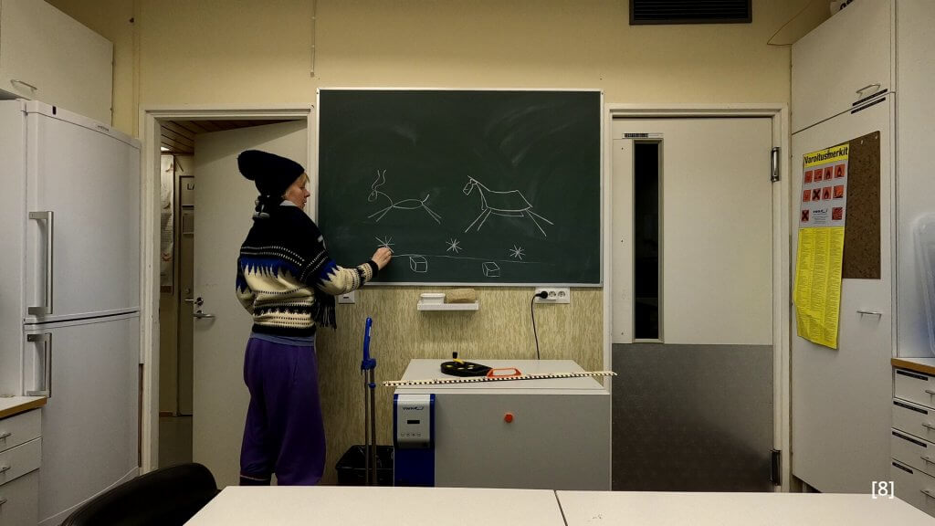 Photograph. Kaisu stands in front of a chalkboard in a classroom. She is drawing reindeer and the permafrost structures beneath them.