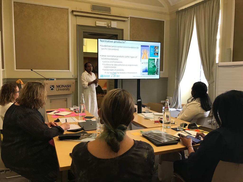 Photo shows a woman presenting to a small group of researchers in a classroom.